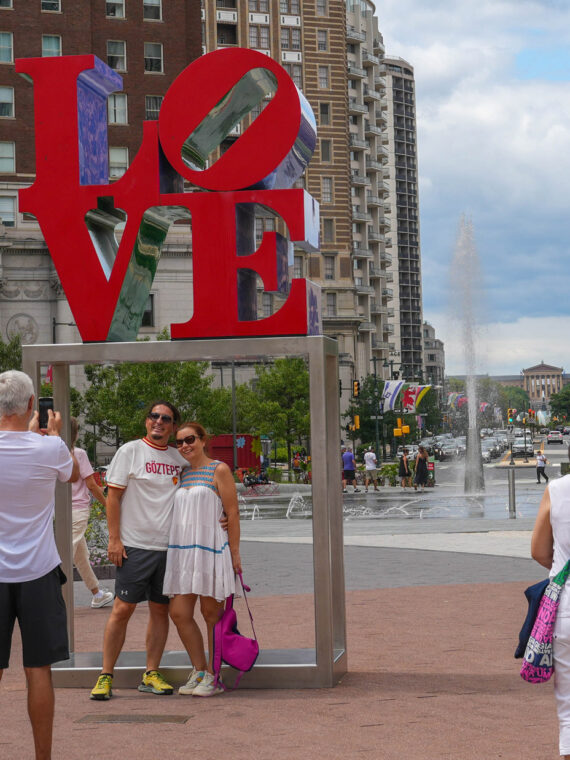 Visitors take photos at the iconic red LOVE sculpture in Philadelphia's Love Park. The large letters are stacked with blue and purple elements visible through the "O" and "E." Historic brick and modern high-rise buildings surround the plaza, with green landscaping, a fountain, and red chairs visible in the background on a partly cloudy day.