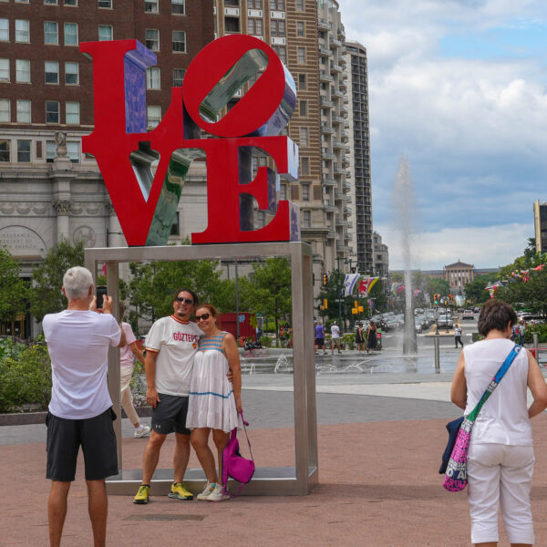 Des visiteurs prennent des photos de l'emblématique sculpture rouge LOVE dans le Love Park de Philadelphie. Les grandes lettres sont empilées et des éléments bleus et violets sont visibles à travers le "O" et le "E". Des bâtiments historiques en brique et des tours modernes entourent la place, avec un aménagement paysager vert, une fontaine et des chaises rouges visibles à l'arrière-plan par temps partiellement nuageux.