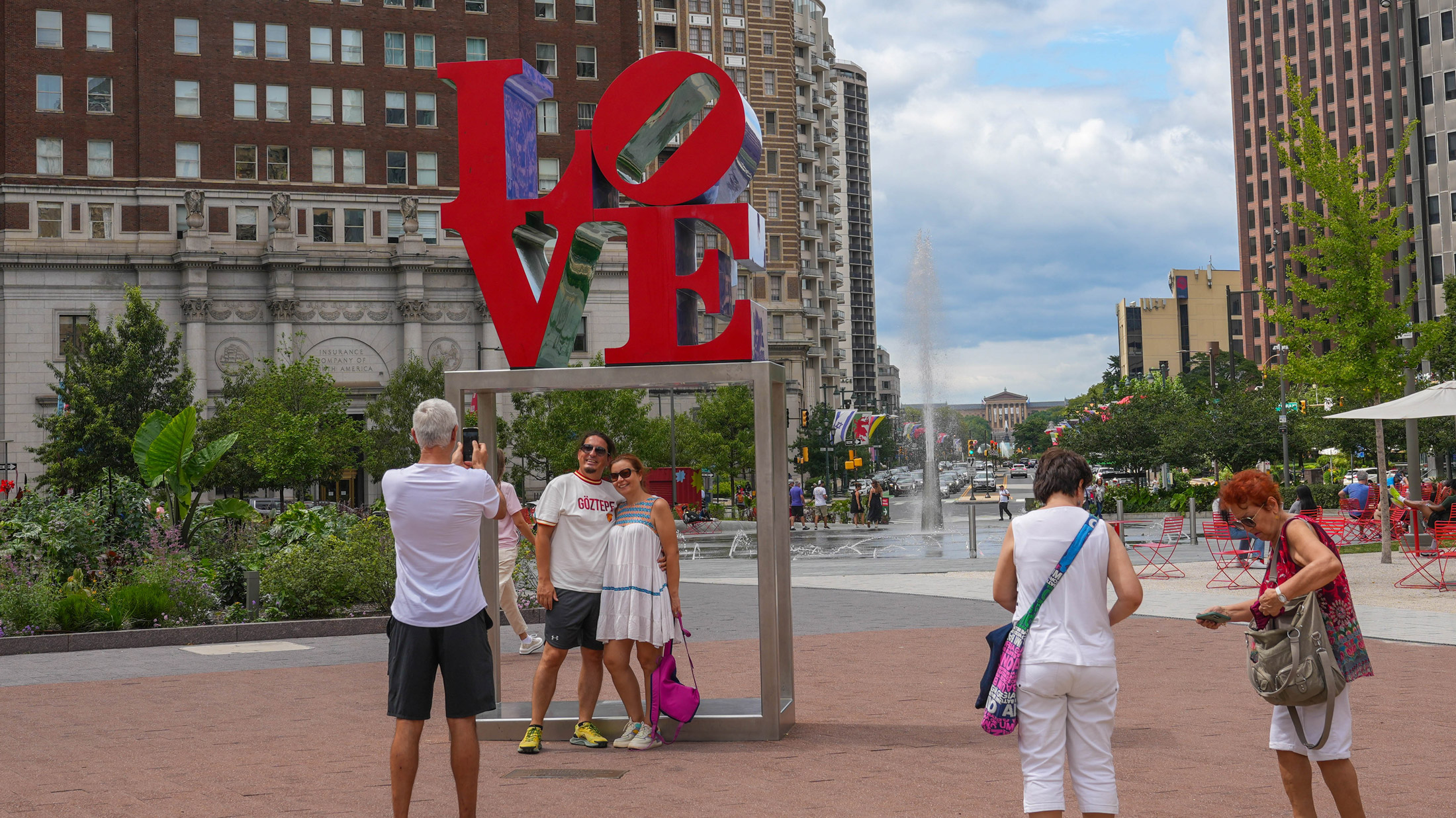 Visitors take photos at the iconic red LOVE sculpture in Philadelphia's Love Park. The large letters are stacked with blue and purple elements visible through the "O" and "E." Historic brick and modern high-rise buildings surround the plaza, with green landscaping, a fountain, and red chairs visible in the background on a partly cloudy day.