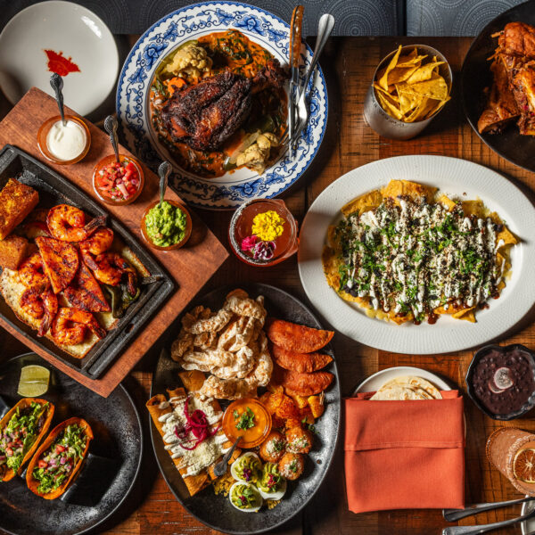 An overhead view of a vibrant Mexican food spread from Mi Vida restaurant featuring multiple dishes arranged on a wooden table. Visible items include grilled meat with charred edges, crispy churros, loaded nachos topped with cheese and herbs, roasted sweet potatoes, fresh salsas in small bowls, guacamole, tacos in black plates, enchiladas on a white plate, fresh lime wedges, and various sauces.