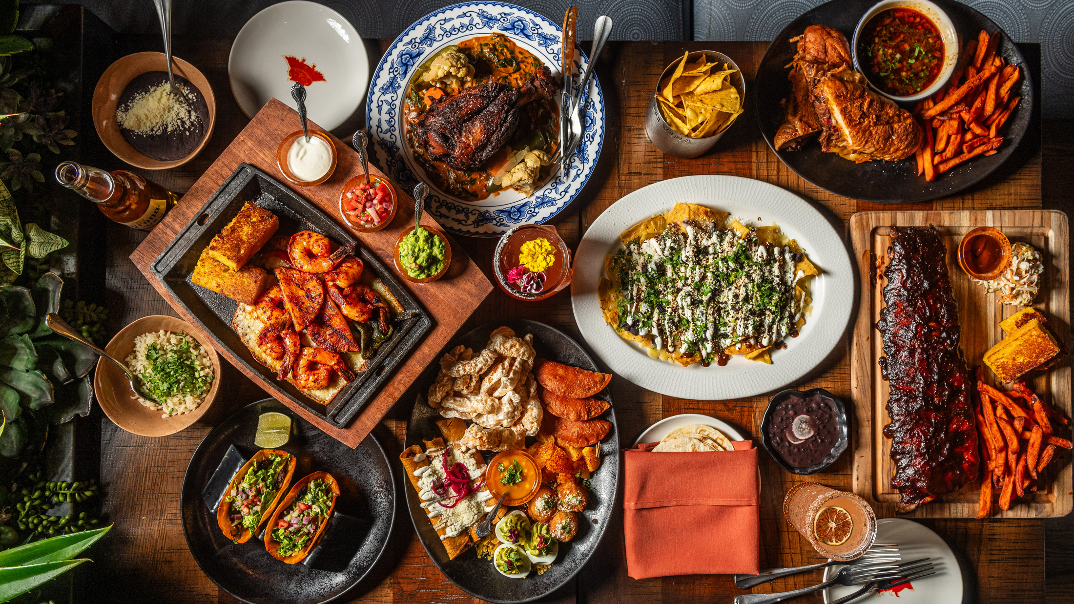 An overhead view of a vibrant Mexican food spread from Mi Vida restaurant featuring multiple dishes arranged on a wooden table. Visible items include grilled meat with charred edges, crispy churros, loaded nachos topped with cheese and herbs, roasted sweet potatoes, fresh salsas in small bowls, guacamole, tacos in black plates, enchiladas on a white plate, fresh lime wedges, and various sauces.