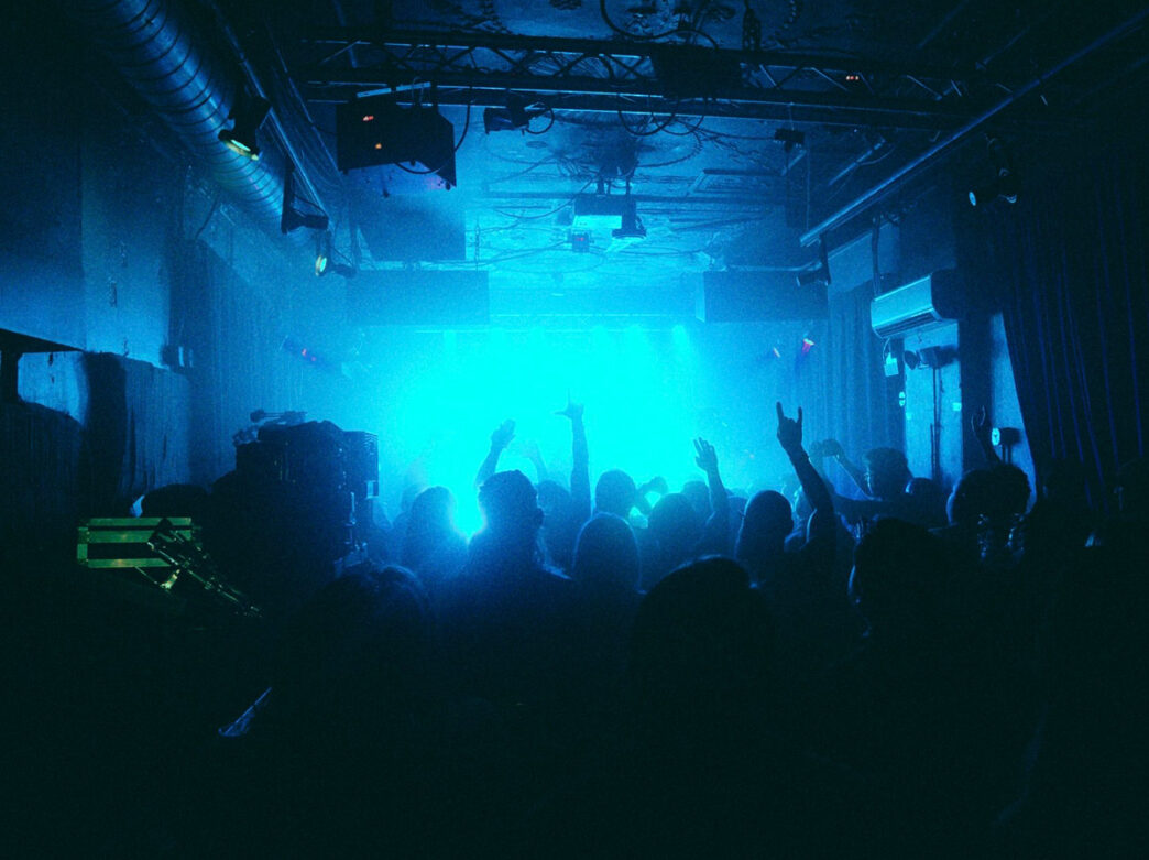 Une foule de spectateurs remplit l'intérieur sombre du MilkBoy à Philadelphie, silhouettée par les lumières bleues de la scène, avec plusieurs membres du public qui lèvent la main pendant une performance en direct.