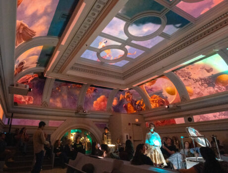 Un grupo de visitantes se reúne en el ornamentado salón del Ministry of Awe, en Filadelfia, y contempla una instalación inmersiva en el techo. El histórico techo con vigas muestra vibrantes imágenes proyectadas en tonos naranjas, rosados, morados y azules, creando una experiencia visual impresionante. Los detalles arquitectónicos incluyen molduras decorativas y puertas en arco, mientras que la iluminación ambiental realza la atmósfera contemplativa.