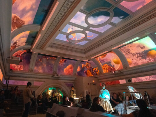 Un groupe de visiteurs se réunit dans le hall orné du Ministry of Awe à Philadelphie, regardant vers le haut une installation immersive au plafond. Le plafond historique à poutres apparentes affiche des images vibrantes projetées en orange, rose, violet et bleu, créant une expérience visuelle impressionnante. Les détails architecturaux comprennent des moulures décoratives et des portes cintrées, tandis que l'éclairage ambiant renforce l'atmosphère contemplative.