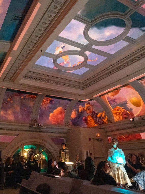 A group of visitors gather in the ornate Hall at Ministry of Awe in Philadelphia, gazing upward at an immersive ceiling installation. The historic beamed ceiling displays vibrant projected imagery in oranges, pinks, purples, and blues, creating an awe-inspiring visual experience. Architectural details include decorative molding and arched doorways, while ambient lighting enhances the contemplative atmosphere.