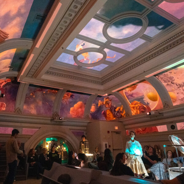 A group of visitors gather in the ornate Hall at Ministry of Awe in Philadelphia, gazing upward at an immersive ceiling installation. The historic beamed ceiling displays vibrant projected imagery in oranges, pinks, purples, and blues, creating an awe-inspiring visual experience. Architectural details include decorative molding and arched doorways, while ambient lighting enhances the contemplative atmosphere.