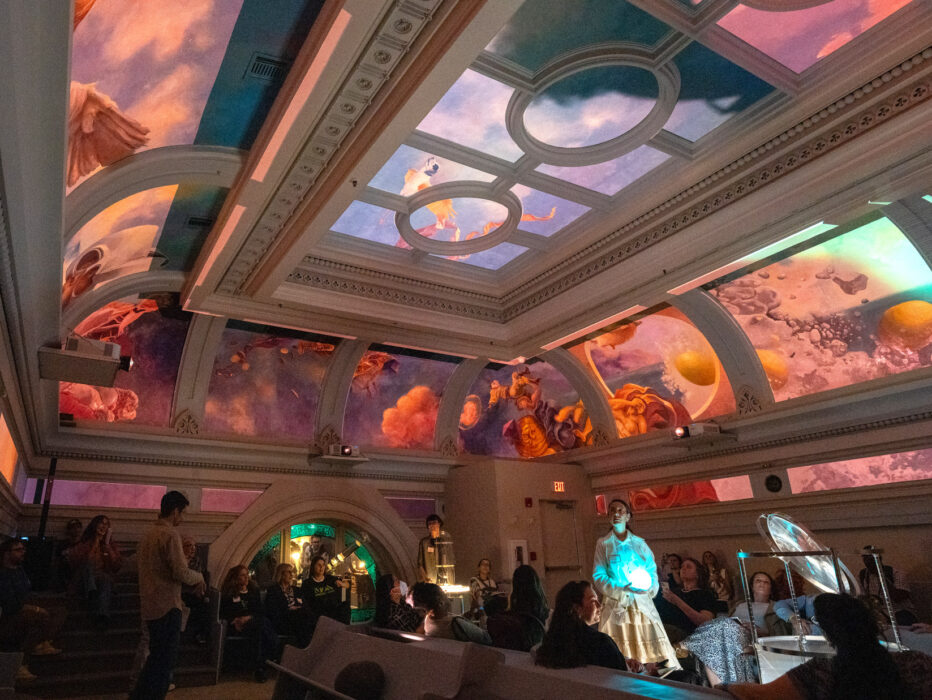 A group of visitors gather in the ornate Hall at Ministry of Awe in Philadelphia, gazing upward at an immersive ceiling installation. The historic beamed ceiling displays vibrant projected imagery in oranges, pinks, purples, and blues, creating an awe-inspiring visual experience. Architectural details include decorative molding and arched doorways, while ambient lighting enhances the contemplative atmosphere.
