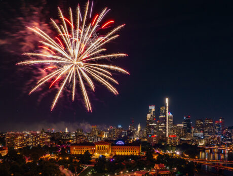 Fireworks burst over the Benjamin Franklin Parkway and Center City Philadelphia skyline during a Fourth of July celebration at night. The illuminated Philadelphia Museum of Art and city skyscrapers are visible beneath colorful fireworks lighting up the sky.