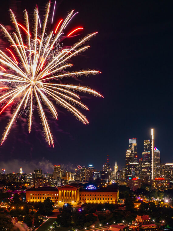 Fireworks burst over the Benjamin Franklin Parkway and Center City Philadelphia skyline during a Fourth of July celebration at night. The illuminated Philadelphia Museum of Art and city skyscrapers are visible beneath colorful fireworks lighting up the sky.