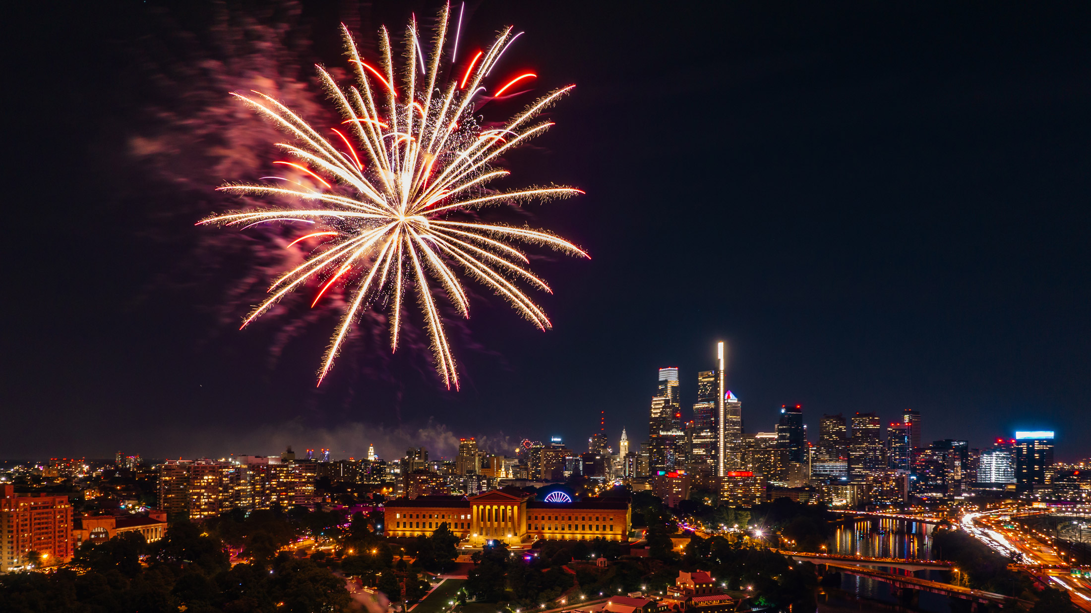 Fireworks burst over the Benjamin Franklin Parkway and Center City Philadelphia skyline during a Fourth of July celebration at night. The illuminated Philadelphia Museum of Art and city skyscrapers are visible beneath colorful fireworks lighting up the sky.
