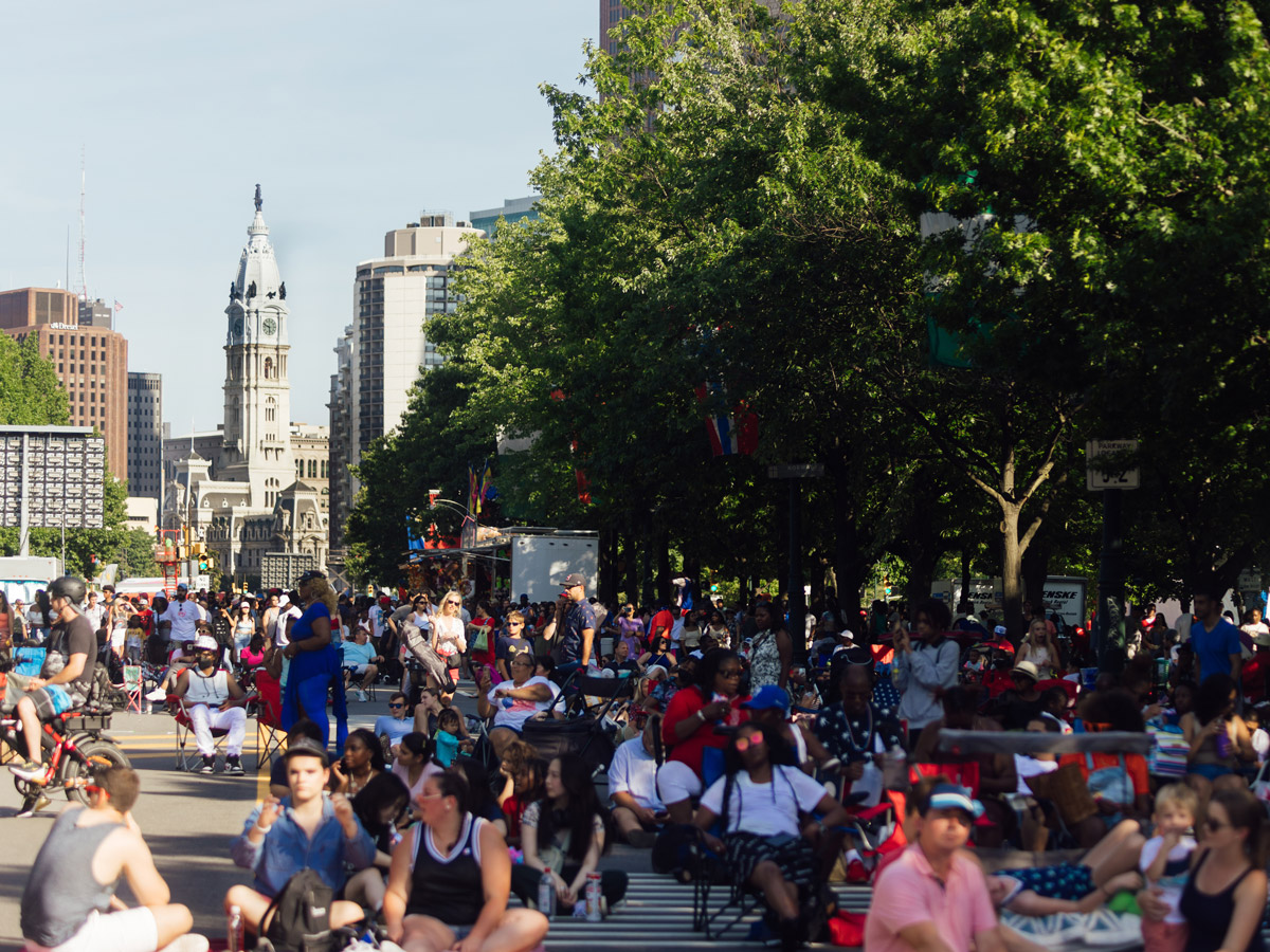 Una multitud se congrega a lo largo de la avenida Benjamin Franklin Parkway en Filadelfia durante la celebración del «Fourth of July Party on the Parkway». La gente se sienta y pasea bajo las calles arboladas, con el Philadelphia City Hall visible al fondo en un soleado día de verano.