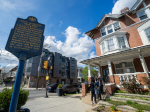 The exterior of the Paul Robeson House in West Philadelphia, a red brick historic rowhouse with white trim and a covered entrance porch. A blue historical marker on the left reads 'Paul Robeson (1898-1976),' describing him as a Rutgers athlete and Columbia law graduate who became a renowned singer and actor. Visitors stand on the front steps of the house. Modern apartment buildings are visible across the street, and the scene is set on a sunny day with blue sky and white clouds.