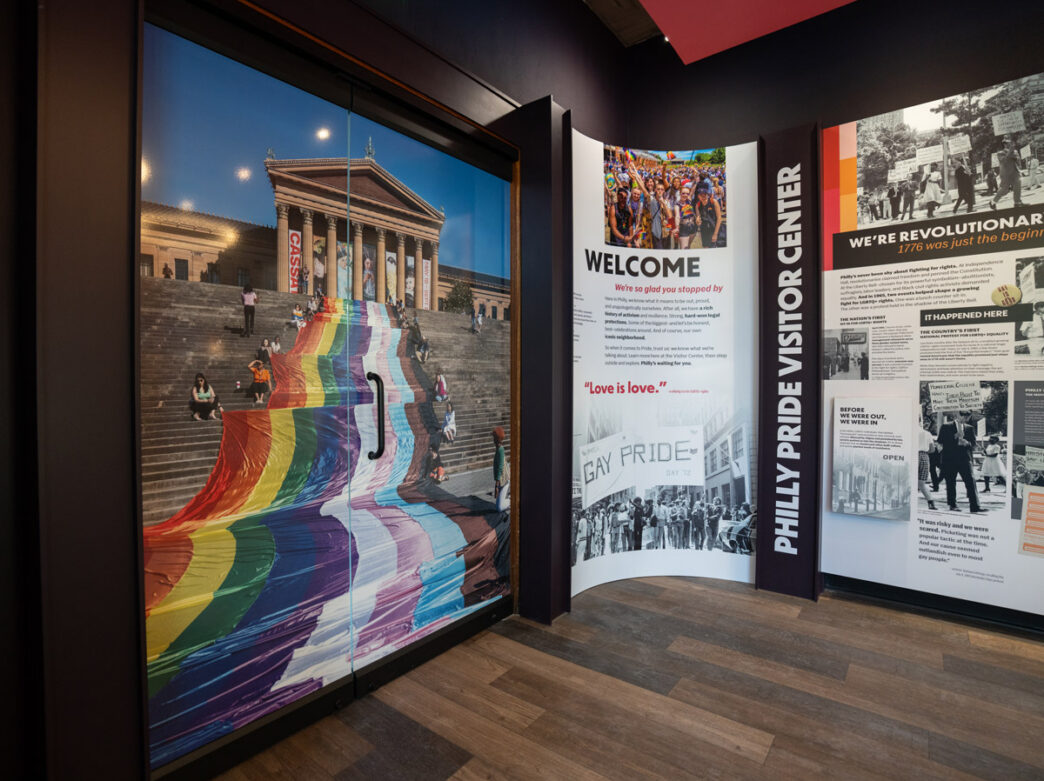 A wall with a large photo of a Pride flag banner on the Philadelphia Museum of Art steps with informational displays showing Philadelphia's role in the fight for LGBTQ+ rights at the Philly Pride Visitor Center.
