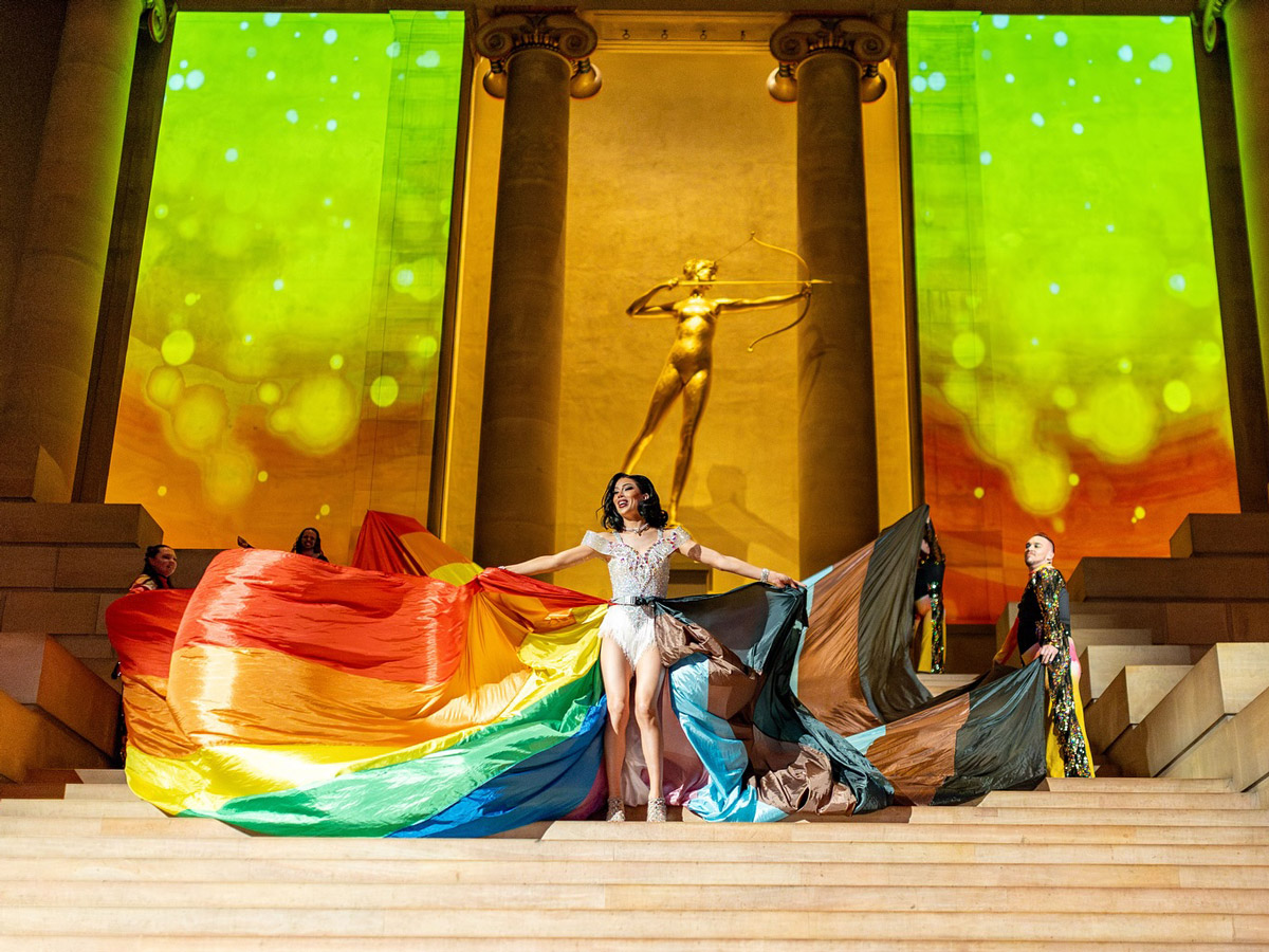 Un artista posa en las escaleras del Philadelphia Museum of Art durante la gala ’Promenade & L.U.V. Awards» de PhillyPride365, sosteniendo una gran bandera arcoíris junto a otros artistas. El evento nocturno cuenta con coloridas proyecciones de luz y la estatua dorada de Diana del museo como telón de fondo.