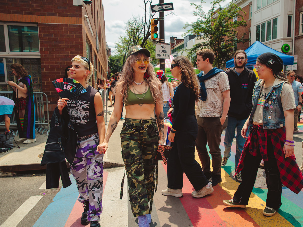 La gente cruza un paso de peatones pintado con los colores del arcoíris durante el Pride Fest Philly en Filadelfia. En primer plano, dos asistentes se dan la mano y sonríen mientras los asistentes al festival se reúnen a lo largo de una calle soleada de la ciudad, decorada con los colores del orgullo y llena de celebraciones comunitarias.