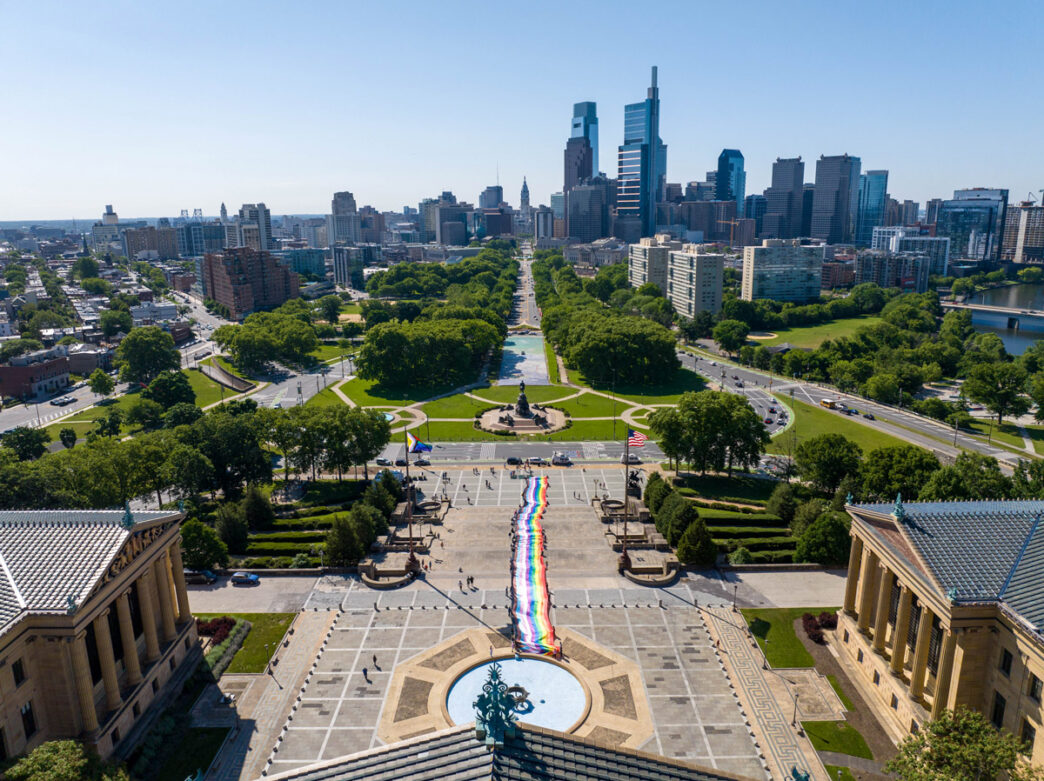 Aerial view of Philadelphia’s Benjamin Franklin Parkway with a long rainbow Pride flag stretching from the Philadelphia Museum of Art toward Center City. The skyline and tree-lined parkway are visible under a clear blue sky.