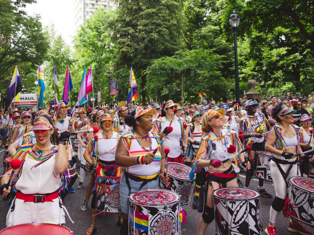 Une batterie colorée se produit pendant la marche des fiertés de Philadelphie. Des musiciens en tenue arc-en-ciel défilent dans une rue bordée d'arbres. La foule se rassemble le long du parcours en agitant des drapeaux de la fierté et en applaudissant cette célébration en plein air.
