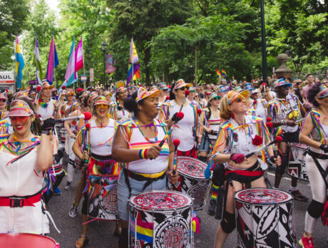 Uma bateria colorida se apresenta durante a Philadelphia Pride March (Marcha do Orgulho da Filadélfia), enquanto músicos em trajes com detalhes de arco-íris marcham por uma rua arborizada. Multidões se reúnem ao longo do percurso, agitando bandeiras do orgulho e aplaudindo durante a comemoração ao ar livre.