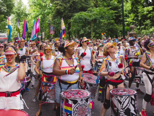 Uma bateria colorida se apresenta durante a Philadelphia Pride March (Marcha do Orgulho da Filadélfia), enquanto músicos em trajes com detalhes de arco-íris marcham por uma rua arborizada. Multidões se reúnem ao longo do percurso, agitando bandeiras do orgulho e aplaudindo durante a comemoração ao ar livre.