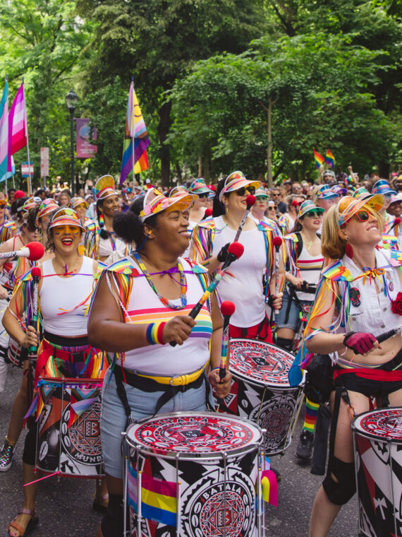Een kleurrijke drumline treedt op tijdens de Philadelphia Pride March terwijl muzikanten in regenboogoutfits door een met bomen omzoomde straat marcheren. Mensenmassa's verzamelen zich langs de route, zwaaien met pride-vlaggen en juichen tijdens de openluchtviering.