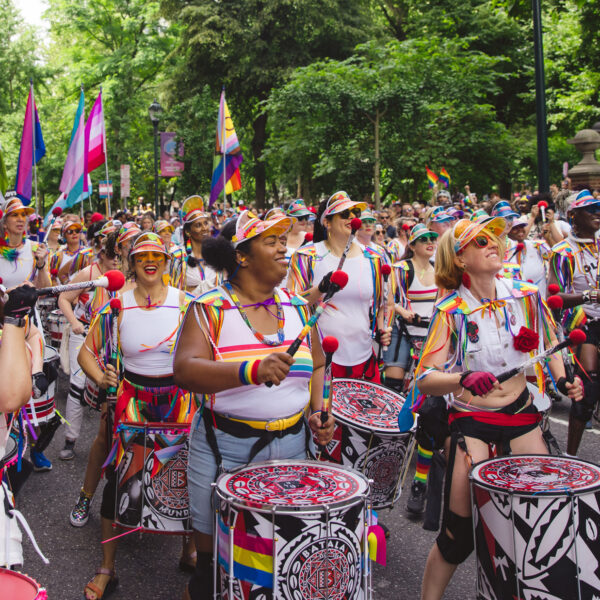 Une batterie colorée se produit pendant la marche des fiertés de Philadelphie. Des musiciens en tenue arc-en-ciel défilent dans une rue bordée d'arbres. La foule se rassemble le long du parcours en agitant des drapeaux de la fierté et en applaudissant cette célébration en plein air.