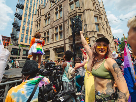 Des personnes font la fête lors d'un festival de rue de la fierté de Philadelphie, tandis qu'un artiste chante sur une scène en plein air. Au premier plan, les participants brandissent des drapeaux arc-en-ciel et des drapeaux de la fierté bisexuelle et dansent devant les bâtiments historiques du centre ville par une journée ensoleillée.