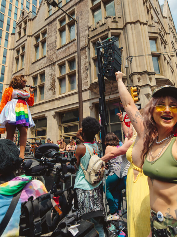 People celebrate at a Philadelphia Pride street festival as a performer sings on an outdoor stage. In the foreground, attendees wave rainbow and bisexual pride flags and dance in front of historic Center City buildings on a sunny day.