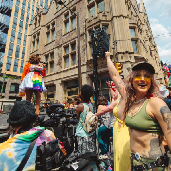 Mensen vieren feest op een Philadelphia Pride straatfestival terwijl een artiest zingt op een buitenpodium. Op de voorgrond zwaaien deelnemers met regenboog- en biseksuele pride-vlaggen en dansen ze voor historische gebouwen in Center City op een zonnige dag.