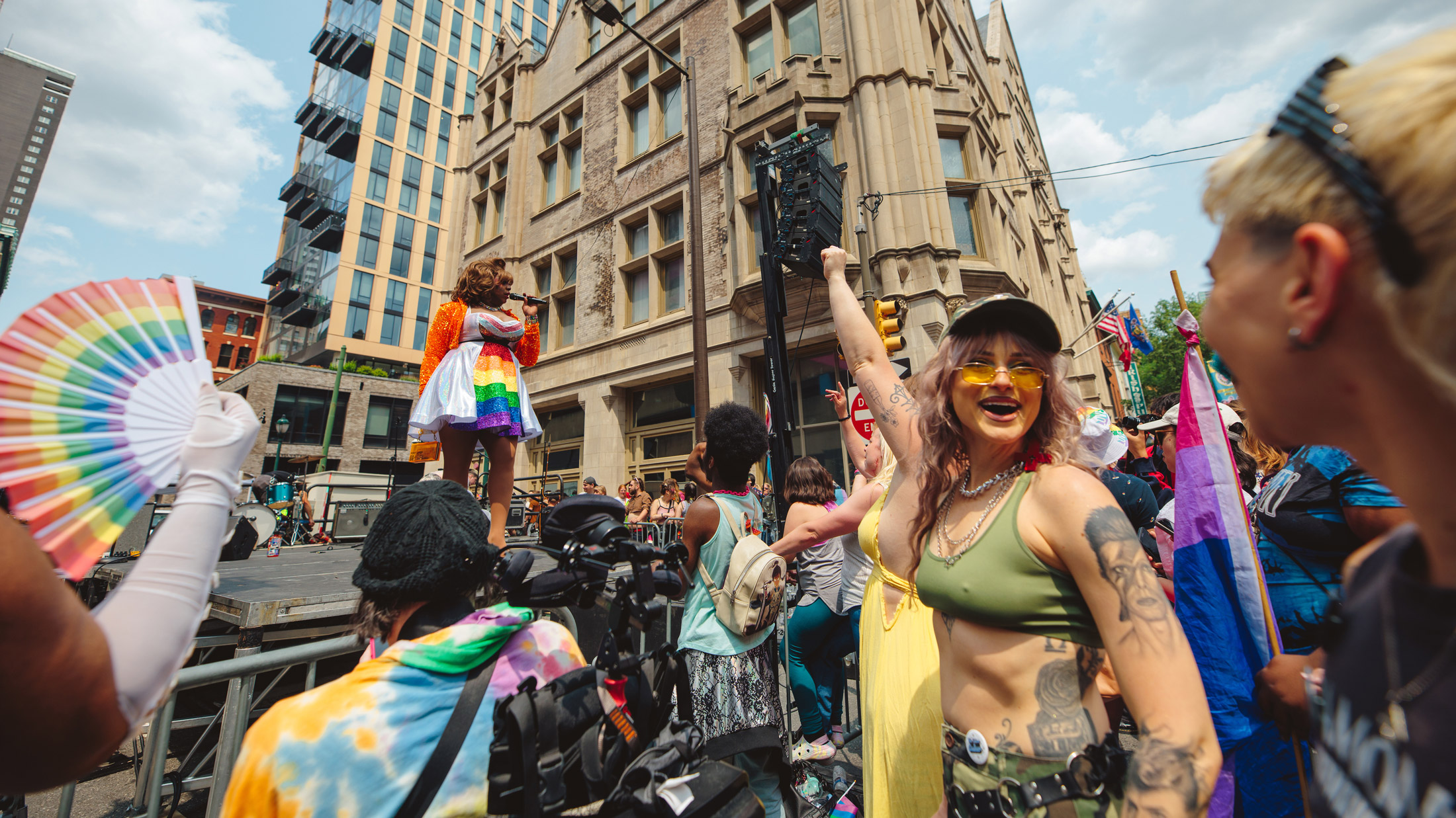 People celebrate at a Philadelphia Pride street festival as a performer sings on an outdoor stage. In the foreground, attendees wave rainbow and bisexual pride flags and dance in front of historic Center City buildings on a sunny day.