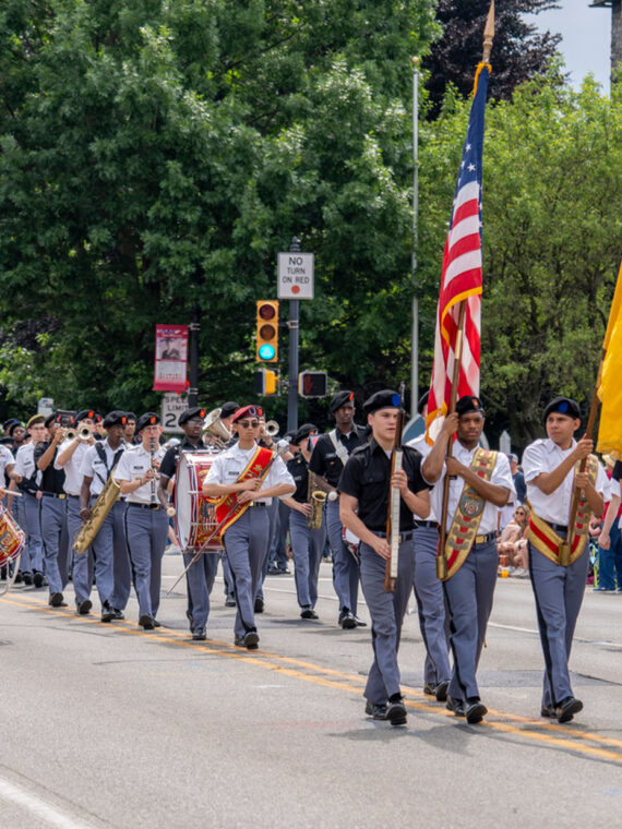 A military marching band in gray uniforms plays brass instruments while marching down a tree-lined street during the Radnor Memorial Day Parade. The band carries American and military flags, marching past crowds of spectators seated along the sidewalk. A stone church building is visible on the right side of the street.