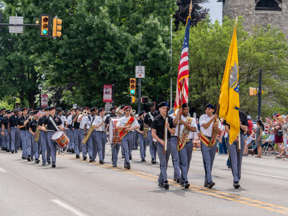 A military marching band in gray uniforms plays brass instruments while marching down a tree-lined street during the Radnor Memorial Day Parade. The band carries American and military flags, marching past crowds of spectators seated along the sidewalk. A stone church building is visible on the right side of the street.