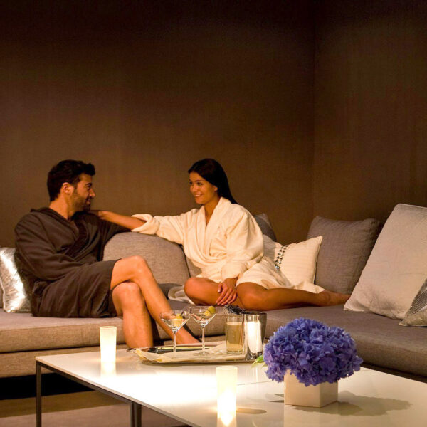 A man and woman sit on a comfortable gray couch in the candlelight. Purple flowers are on the table