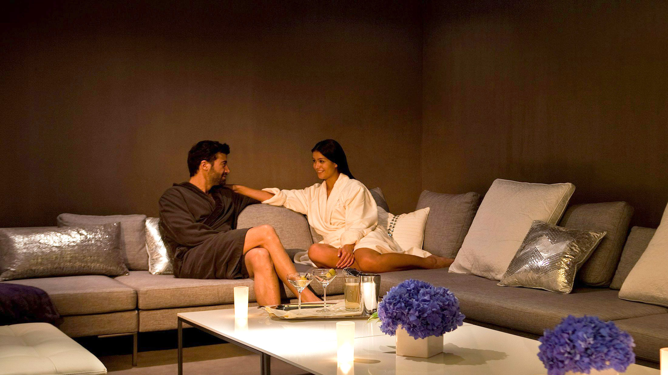 A man and woman sit on a comfortable gray couch in the candlelight. Purple flowers are on the table