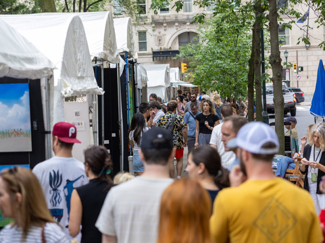 Crowds browse rows of white artist tents at the Rittenhouse Square Fine Art Show in Philadelphia, strolling along the tree-lined sidewalk while viewing paintings and artwork displays.
