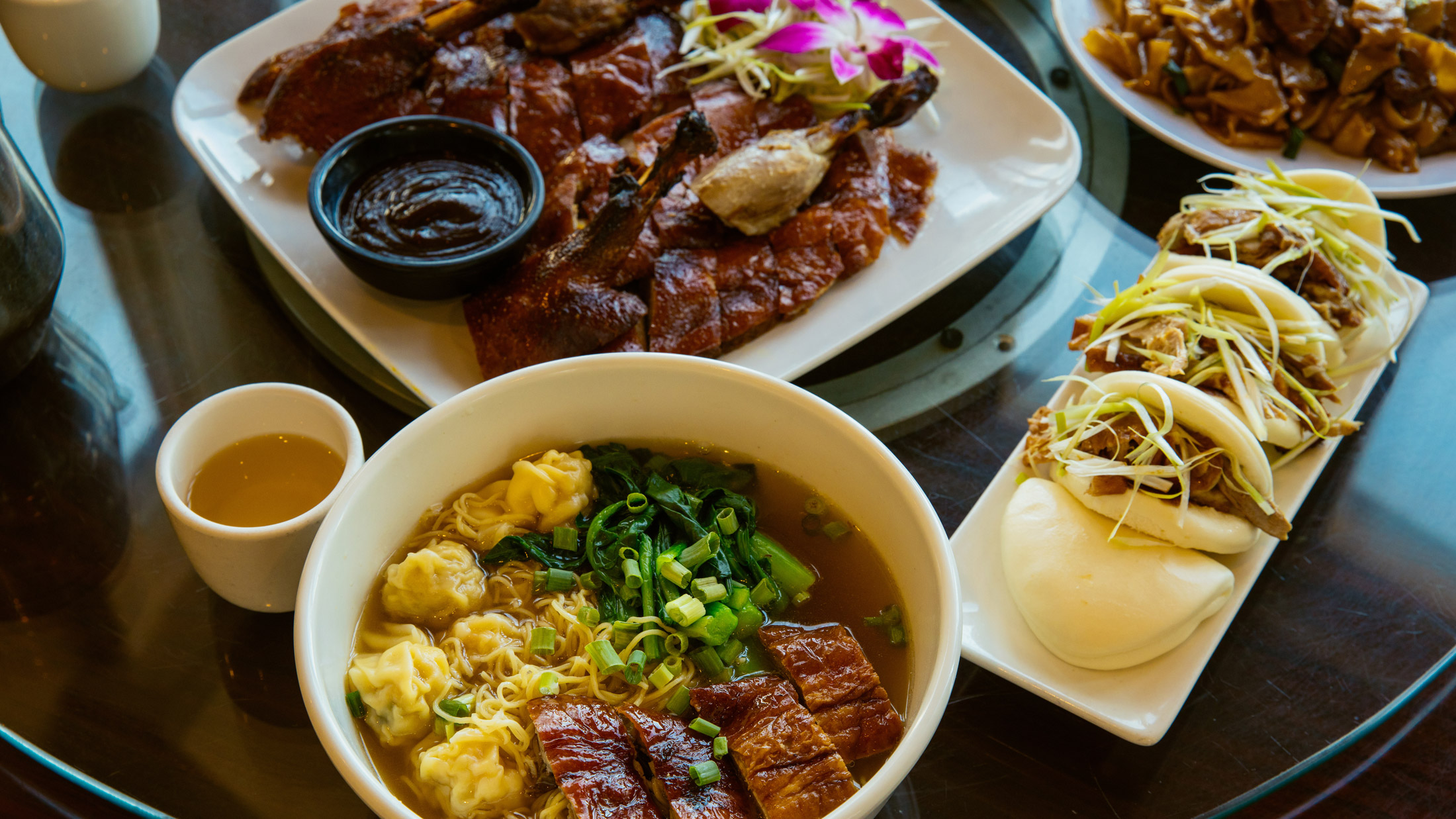 A table full of Asian cuisine, including a plate of roasted duck and a bowl of wonton noodle soup at Sang Kee Peking Duck House