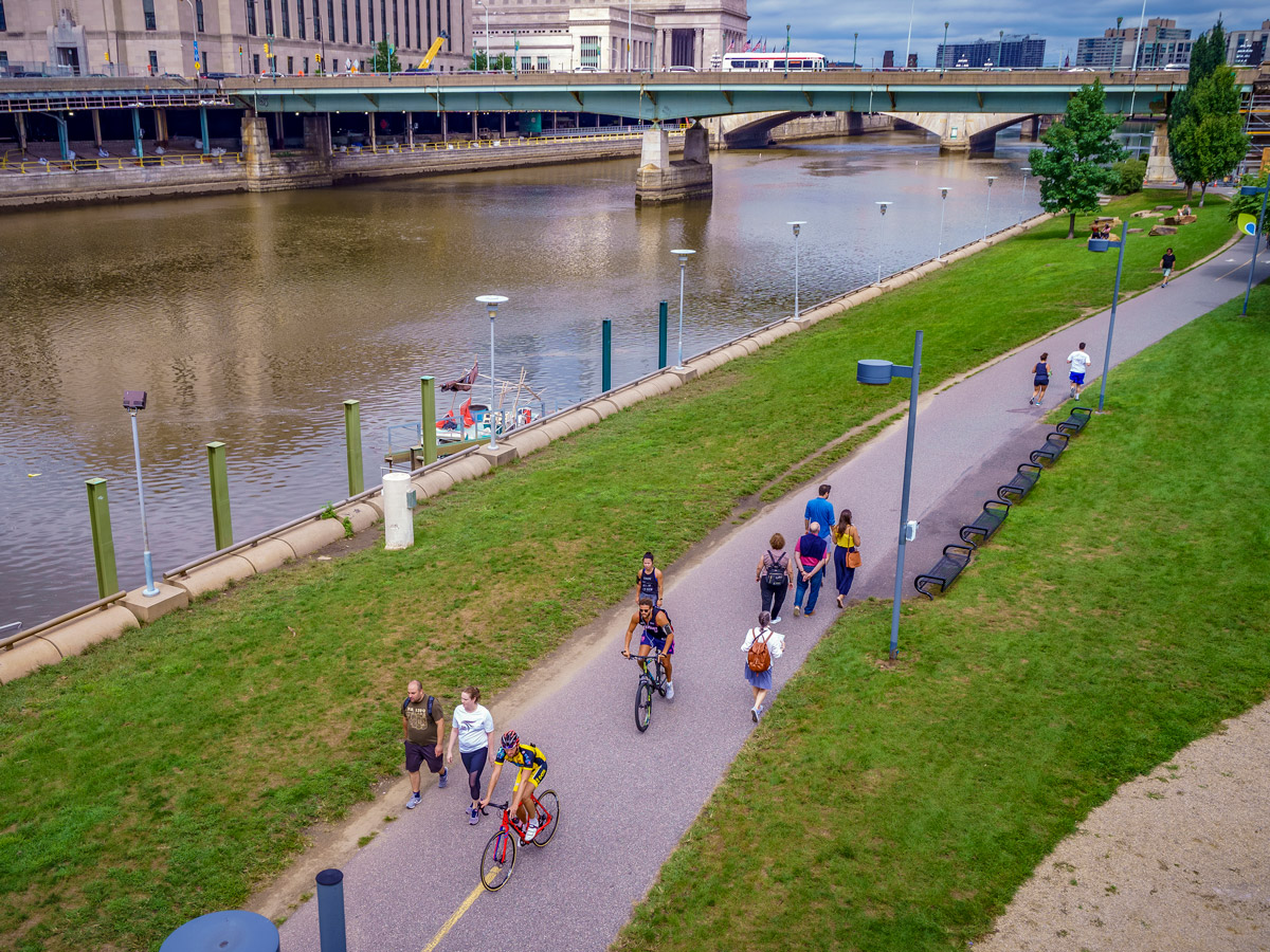 Aerial view of the Schuylkill River Trail in Philadelphia showing cyclists and pedestrians on a paved path alongside green lawns, with the Schuylkill River, multiple bridges, and downtown buildings in the background.