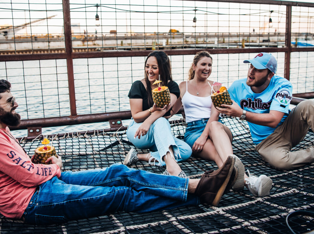 Quatre amis se détendent ensemble sur une aire de repos en filet au Spruce Street Harbor Park à Philadelphie, en riant et en buvant des boissons tropicales à base d'ananas au bord de la rivière Delaware au coucher du soleil. Des guirlandes lumineuses sont suspendues au-dessus de la tête, tandis que les bateaux et le front de mer créent une atmosphère estivale animée en arrière-plan.