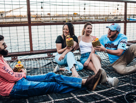 Four friends relax together on a netted lounge area at Spruce Street Harbor Park in Philadelphia, laughing and holding tropical pineapple drinks beside the Delaware River at sunset. String lights hang overhead while boats and the waterfront create a lively summer atmosphere in the background.