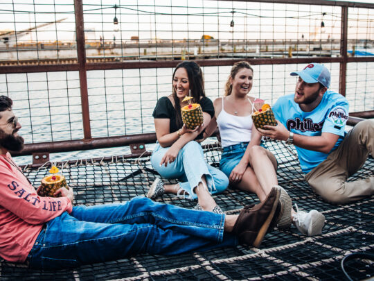 Four friends relax together on a netted lounge area at Spruce Street Harbor Park in Philadelphia, laughing and holding tropical pineapple drinks beside the Delaware River at sunset. String lights hang overhead while boats and the waterfront create a lively summer atmosphere in the background.