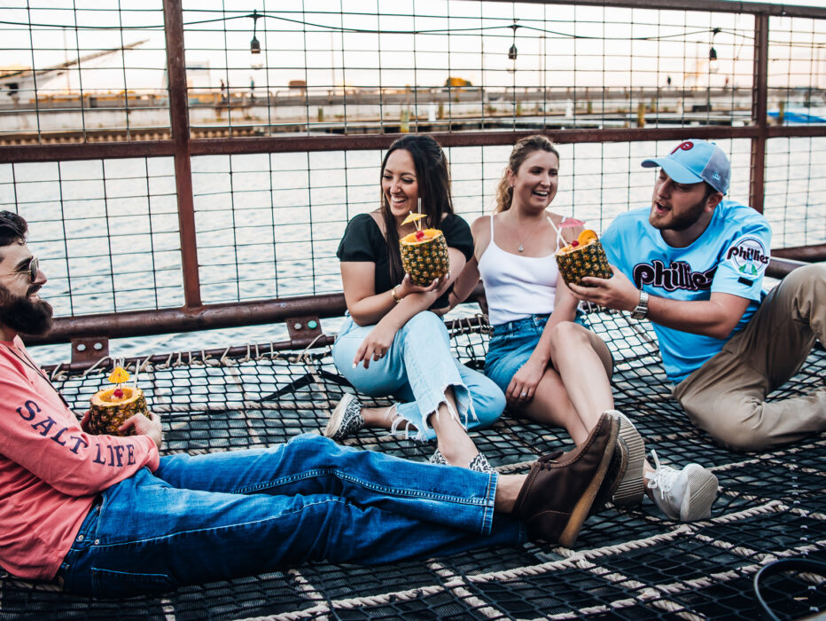 Quatre amis se détendent ensemble sur une aire de repos en filet au Spruce Street Harbor Park à Philadelphie, en riant et en buvant des boissons tropicales à base d'ananas au bord de la rivière Delaware au coucher du soleil. Des guirlandes lumineuses sont suspendues au-dessus de la tête, tandis que les bateaux et le front de mer créent une atmosphère estivale animée en arrière-plan.