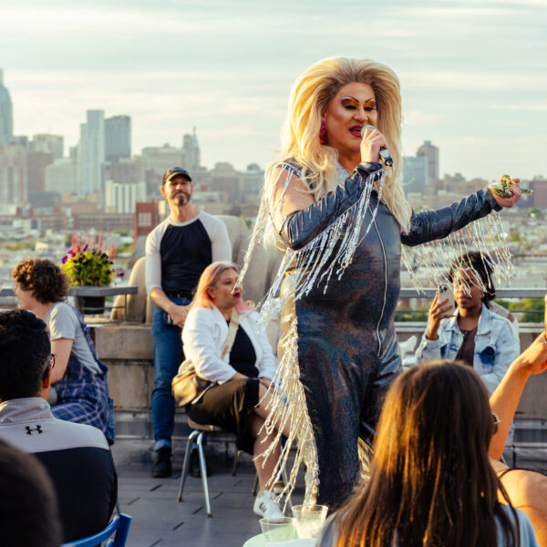 Un artista drag vestido con un traje negro con flecos brillantes habla por un micrófono en el Bok Bar, con el horizonte de Filadelfia como telón de fondo.