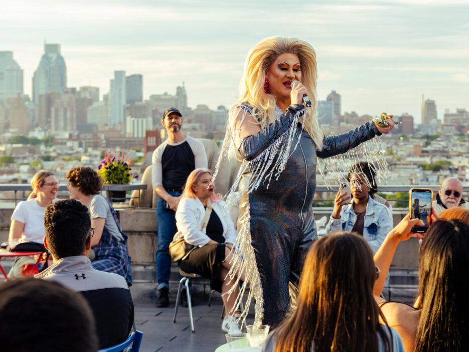 Un artista drag vestido con un traje negro con flecos brillantes habla por un micrófono en el Bok Bar, con el horizonte de Filadelfia como telón de fondo.
