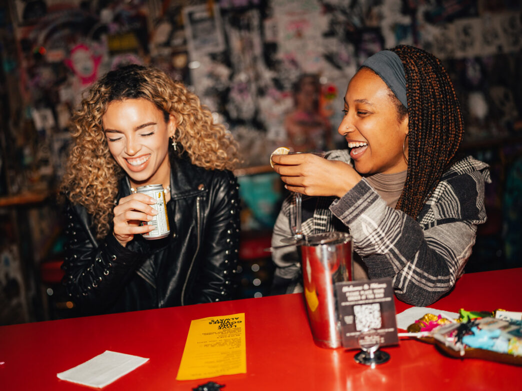 Deux personnes rient et prennent un verre ensemble au bar de Tattooed Mom, à Philadelphie. Le fond coloré, couvert de graffitis, et le bar rouge vif créent une atmosphère vivante et éclectique.