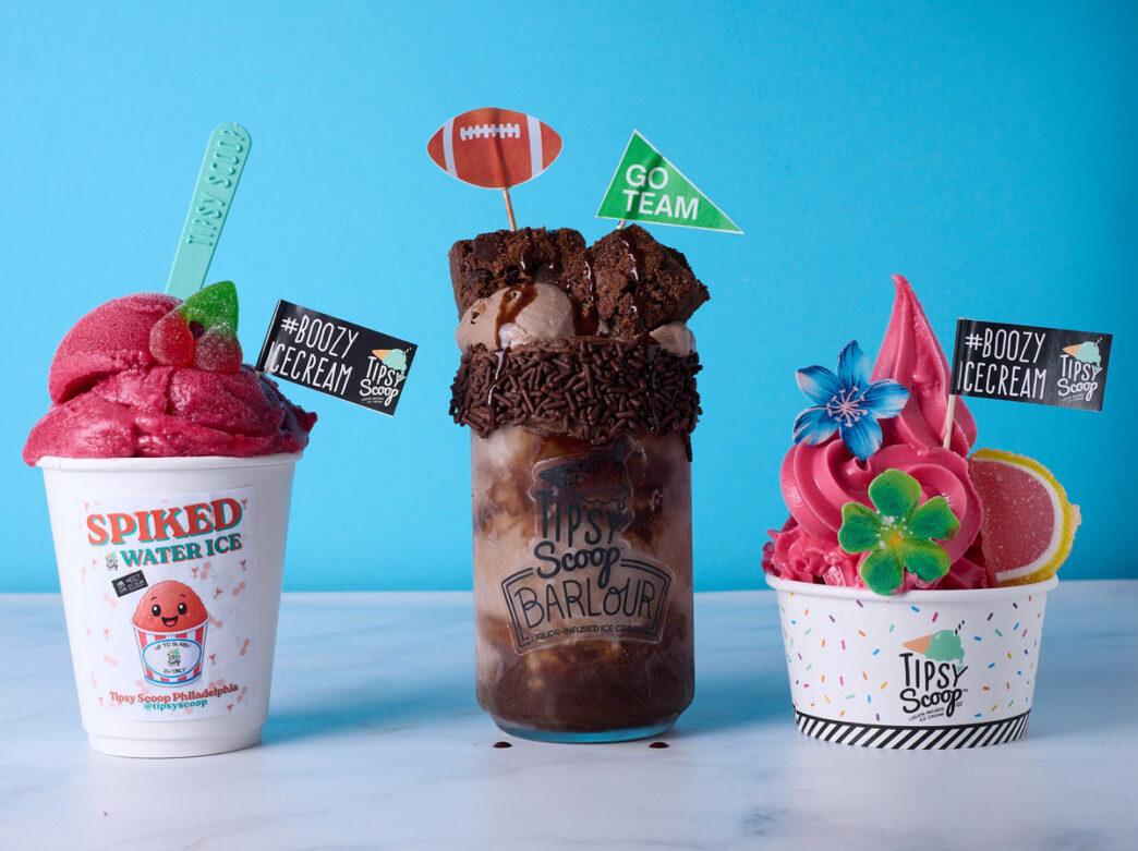 Three colorful alcoholic ice cream desserts from Tipsy Scoop Philadelphia displayed against a bright blue background. On the left is a pink "Spiked Water Ice" in a white cup topped with a cherry and spoon. In the center is a chocolate ice cream sundae topped with chocolate shavings and a football-shaped decoration. On the right is a bright pink ice cream swirl topped with decorative flowers and citrus slices. Each cup features the Tipsy Scoop branding and "#BOOZY ICECREAM" labels.