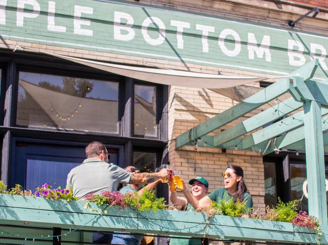 Les gens lèvent leur verre et sourient dans le patio extérieur de Triple Bottom Brewing à Philadelphie, avec son extérieur peint en vert et ses sièges bordés de fleurs, lors d'une journée ensoleillée.