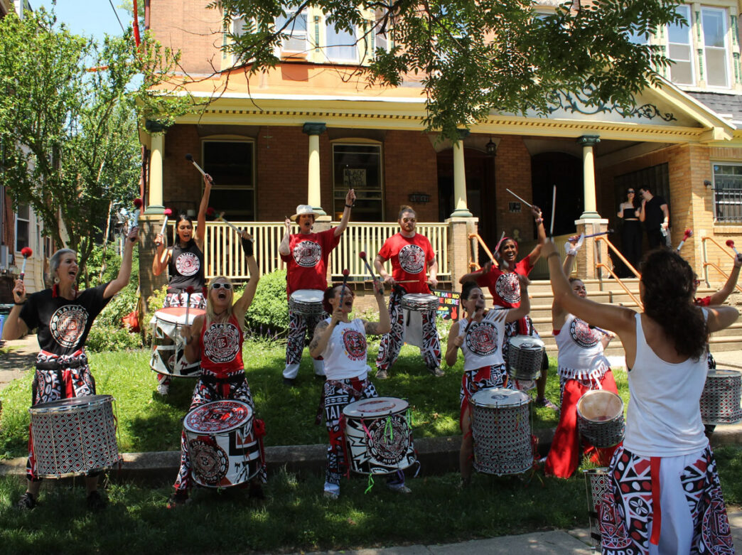 Een levendig drumensemble treedt op in een woonwijk tijdens West Philly Porchfest, met muzikanten in gecoördineerde rode, zwarte en witte outfits die percussie-instrumenten bespelen voor een portiek van een rijtjeshuis.