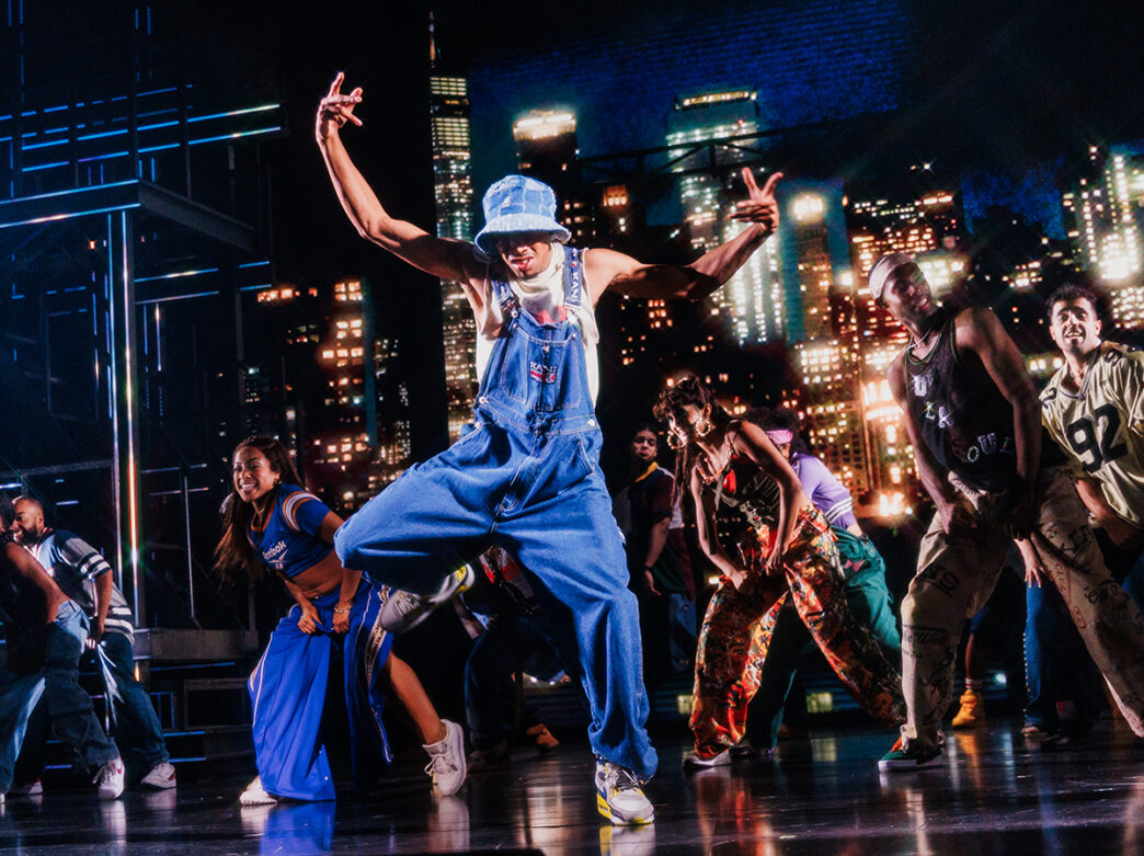 A high-energy cast, including a central dancer wearing overalls and jean bucket hat, performs an electrifying hip-hop dance during the production of Hell's Kitchen.