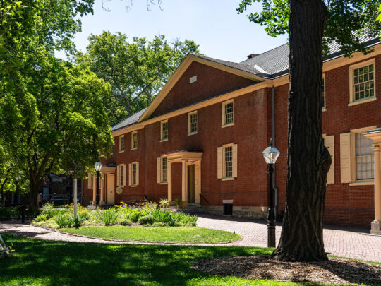 Sunlight filters through mature green trees onto the brick facade of Arch Street Meeting House, with landscaped gardens, brick walkways and historic lampposts in the foreground.