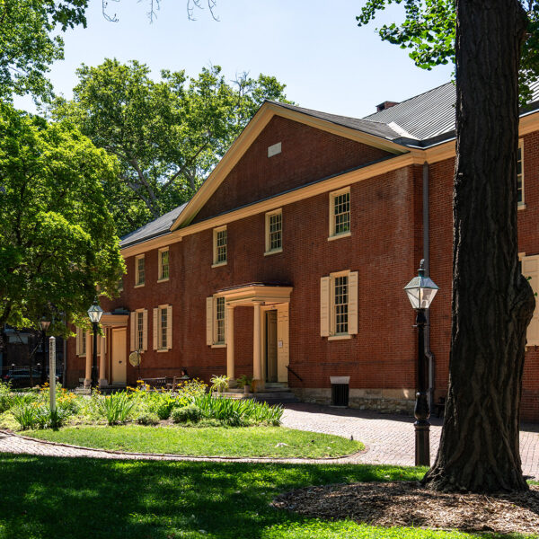 A luz do sol penetra por entre as árvores verdes e maduras e incide sobre a fachada de tijolos da Arch Street Meeting House, com jardins bem cuidados, calçadas de tijolos e postes de luz históricos em primeiro plano.