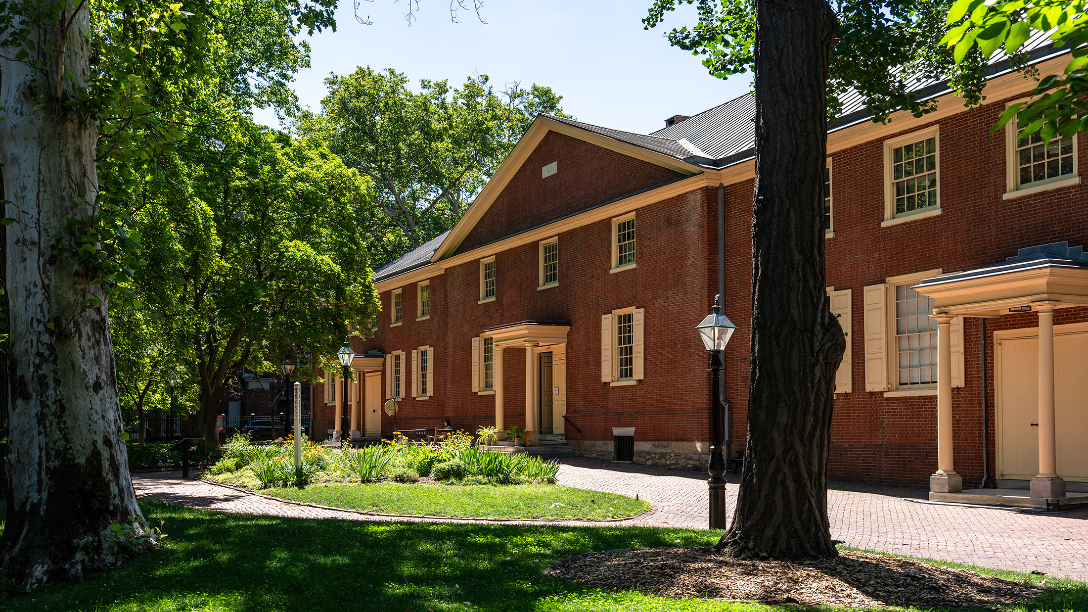 Sunlight filters through mature green trees onto the brick facade of Arch Street Meeting House, with landscaped gardens, brick walkways and historic lampposts in the foreground.